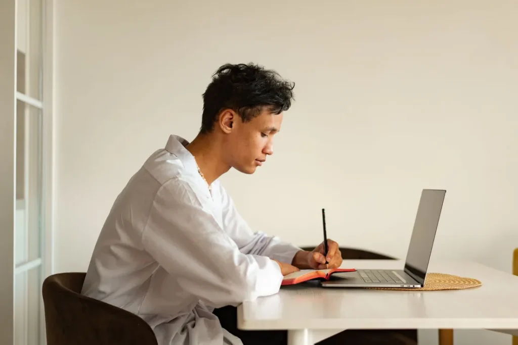 Pessoa estudando em casa com caderno e computador.